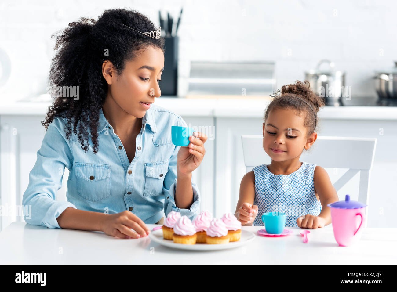 Afrikanische amerikanische Mutter und Tochter mit Tiaras am Tisch sitzen mit Cupcakes in der Küche Stockfoto