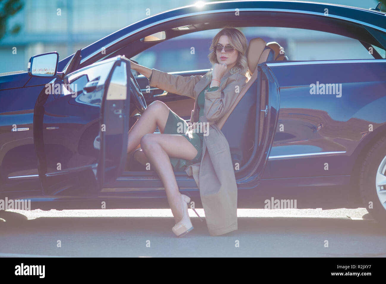 Bild der Blonde mit Brille und langes Kleid und High Heels Schuhe im Auto sitzen mit offenen Tür am Nachmittag. Sonnenlicht Wirkung. Stockfoto