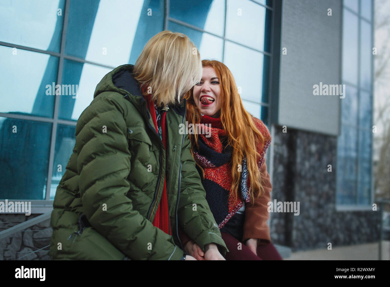 Junge europäische Paar Spaß im Winter im städtischen Hintergrund. Punks oder Hipster Style. Rothaarige Mädchen ist in roten Schal bekleidet, braunen Fell, kurze Shorts und Tights. Blasse Mann in Grün bolognese Jacke bekleidet, roter Schal und Jeans. Stockfoto