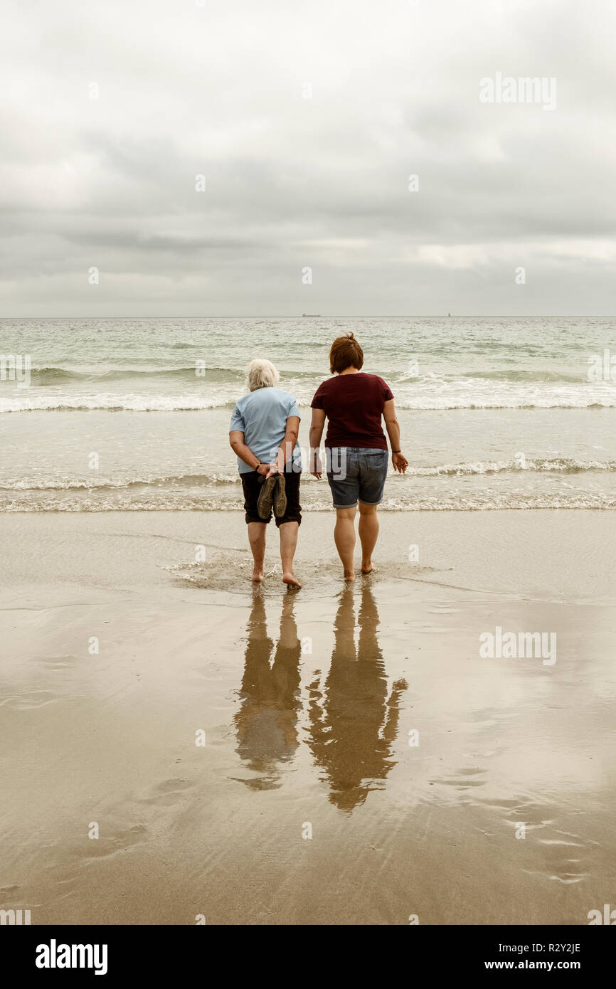 Rückansicht eines grauen Haaren ältere Frau und eine jüngere Frau mit Schuhe aus Paddeln in seichten Wellen auf einem Sandstrand. Stockfoto