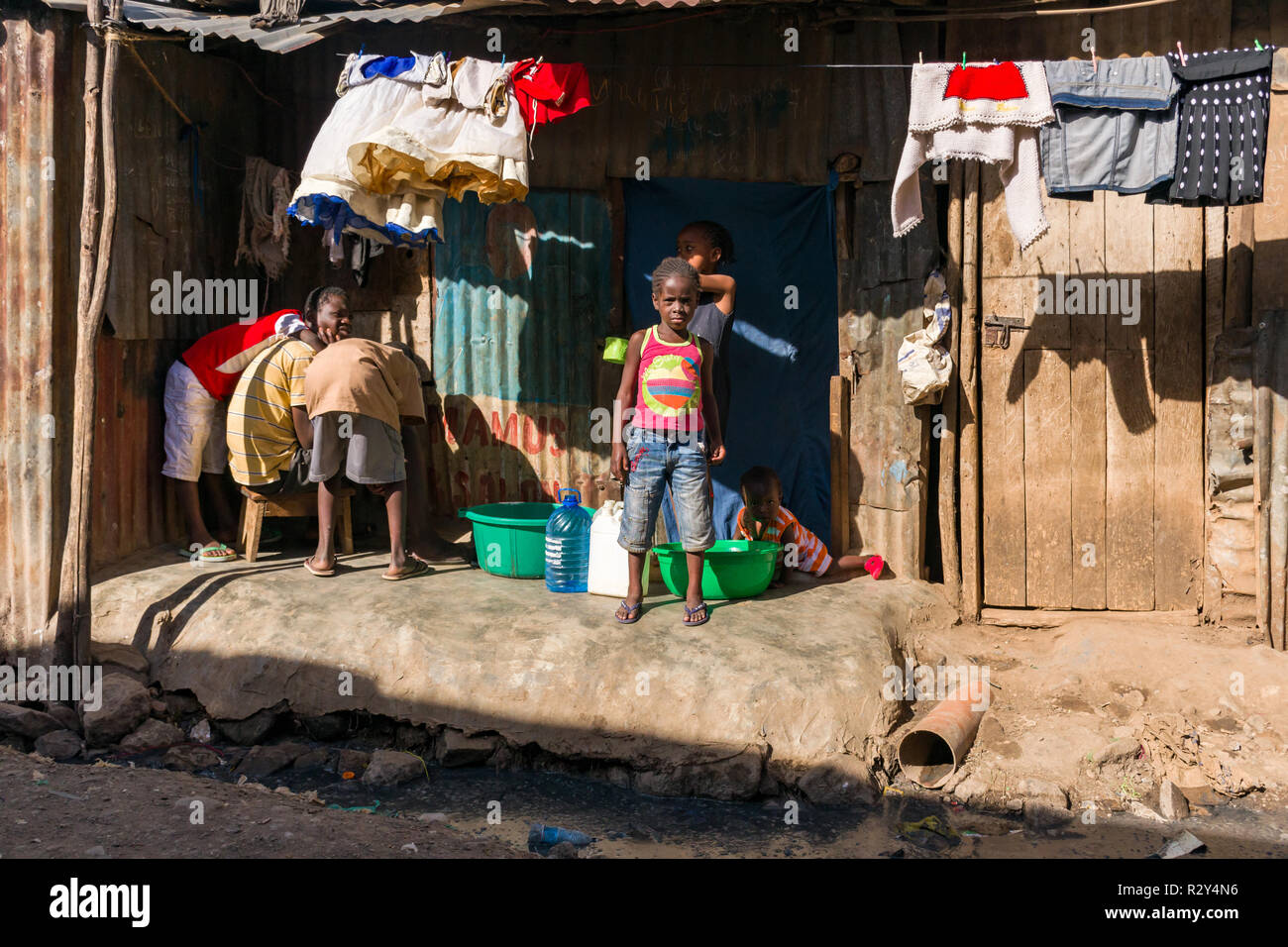 Kinder sitzen außerhalb einer Wellblech Hütte mit Waschmaschine hängen auf einer Linie und offenen Abwasserkanal vor Ihnen, Mathare, Kenia Stockfoto