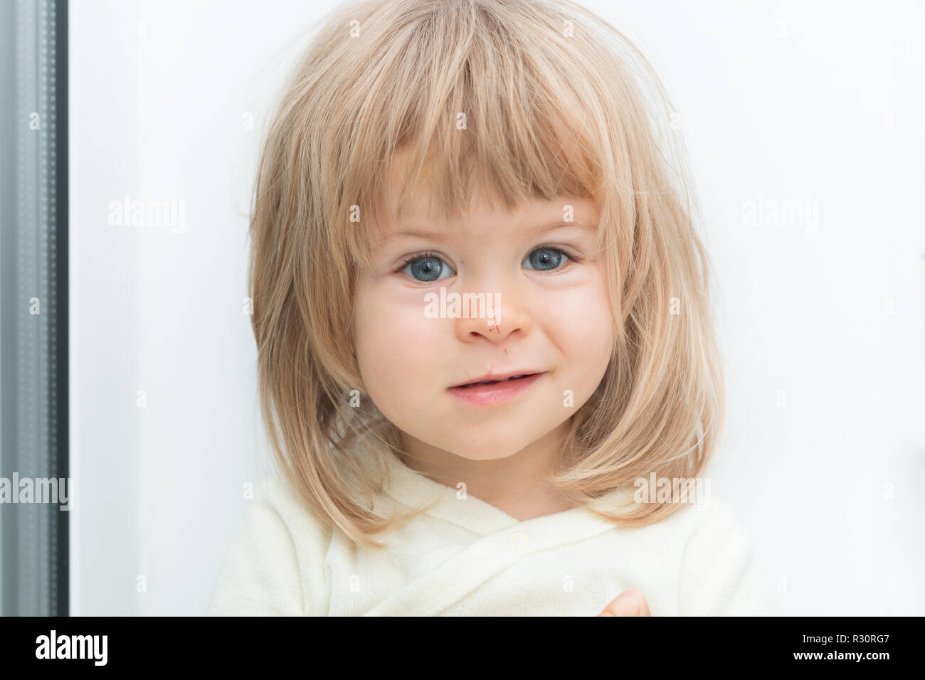 Headshot von adorable cute Blonde weibliches Kind auf Kamera mit schweren umgekippt Gesichtsausdruck. Grinsen auf dem Gesicht. Hübsches kleines Mädchen mit kaukasischen Merkmale und Kratzer auf der Nase. Attraktive baby Stockfoto