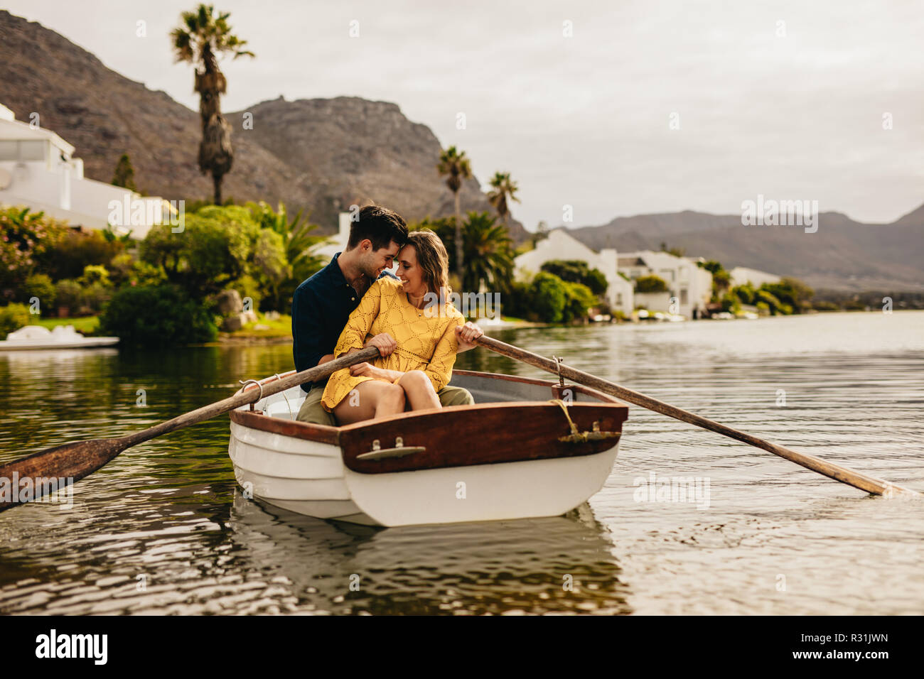 Junges Paar sitzen gemeinsam in einem Boot ihre Köpfe zu berühren. Paar Spaß auf einem Boot Datum in einem See mit Hügeln und Häuser im Hintergrund. Stockfoto