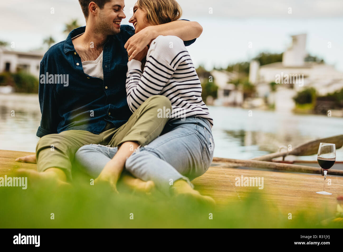 Lächelnden jungen Mann und Frau zusammen auf einer hölzernen Dock in der Nähe von einem See am jeweils anderen suchen. Verliebtes Paar in der Nähe von einem See jedes andere Holding sitzen Stockfoto