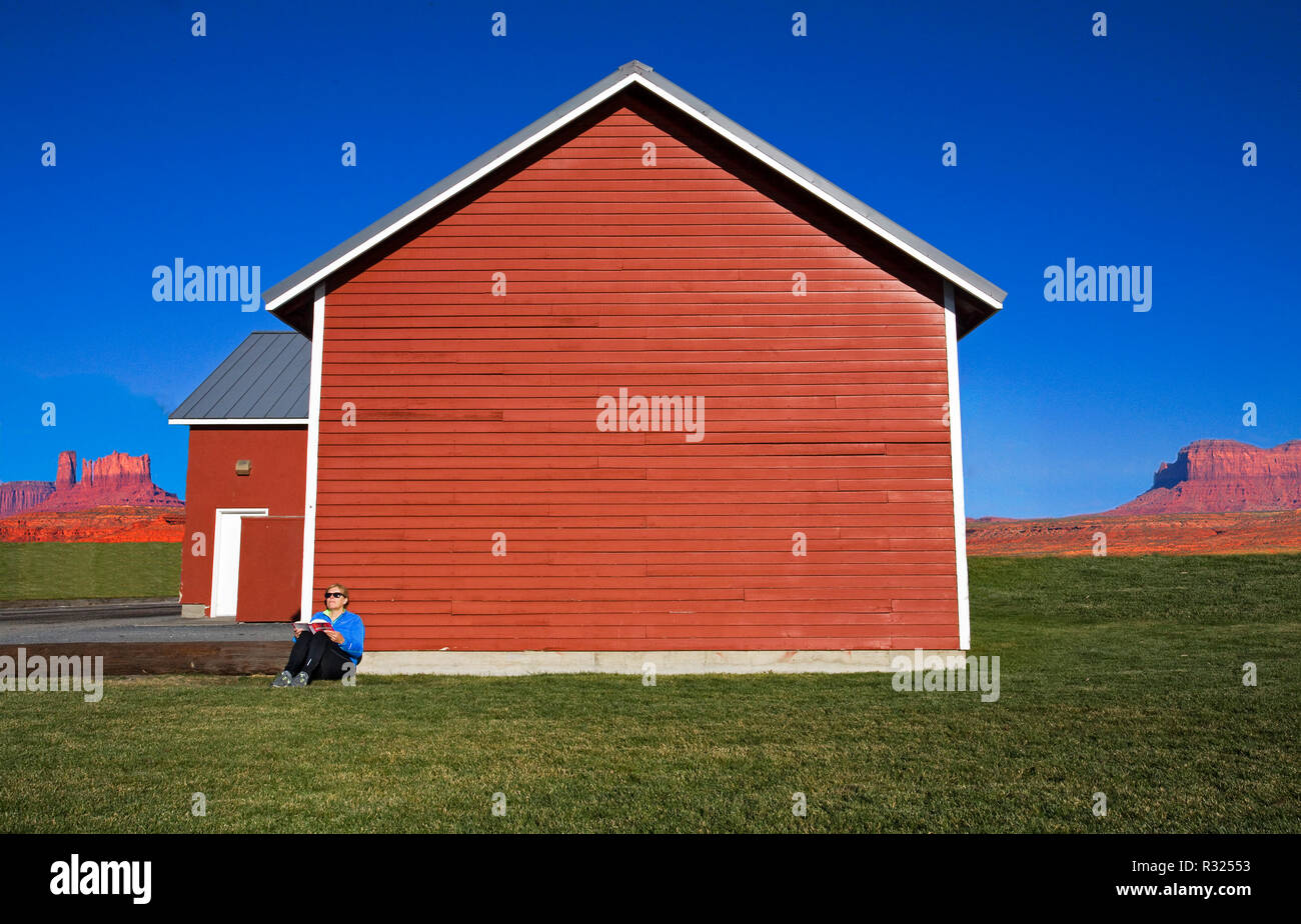 Eine einzelne Frau auf dem Boden sitzend lesen, als sie sich gegen eine rote Holzhaus an einem sonnigen Herbsttag neigt. Stockfoto