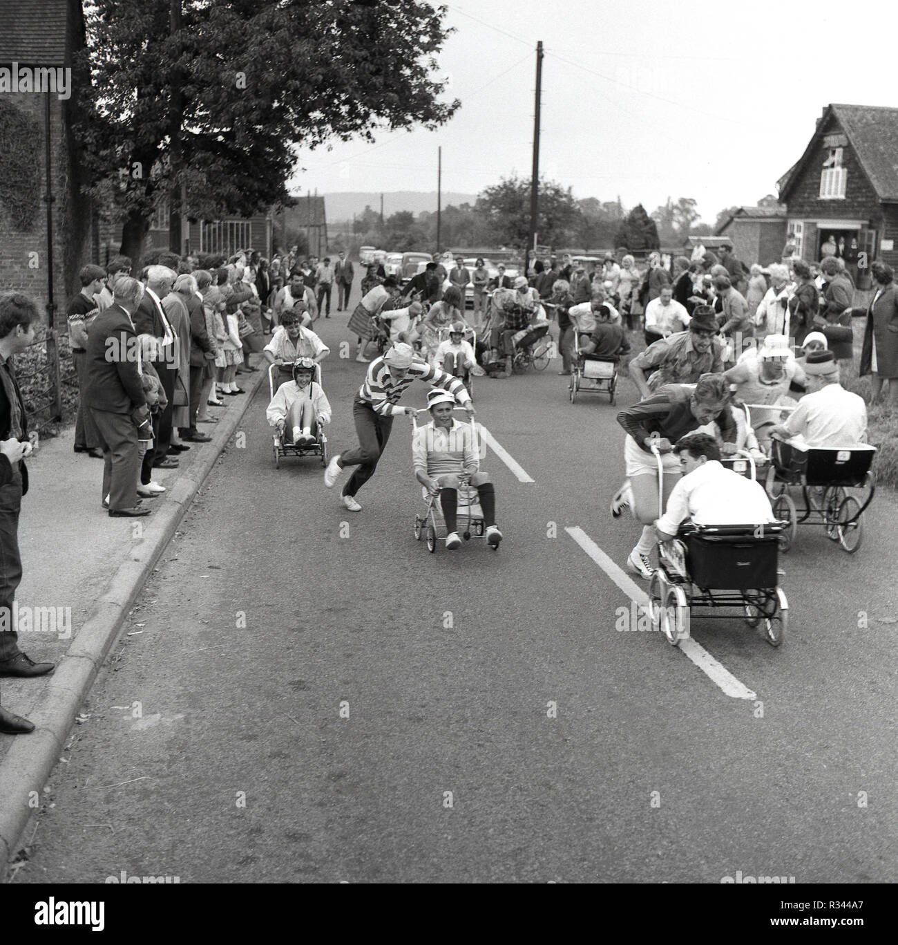 1967, und Ihre aus... der anfang des jährlichen pram Rennen erhält unterwegs bei Quainton Dorf, in der Nähe von Aylesbury, Buckinghamshire, England, UK. Ein eindeutig exzentrischen Britischen Dorf Ereignis, es sieht erwachsene Männer, verkleidet als "Babys im Kinderwagen sitzen und schob mit hoher Geschwindigkeit entlang einer Landstraße. Stockfoto
