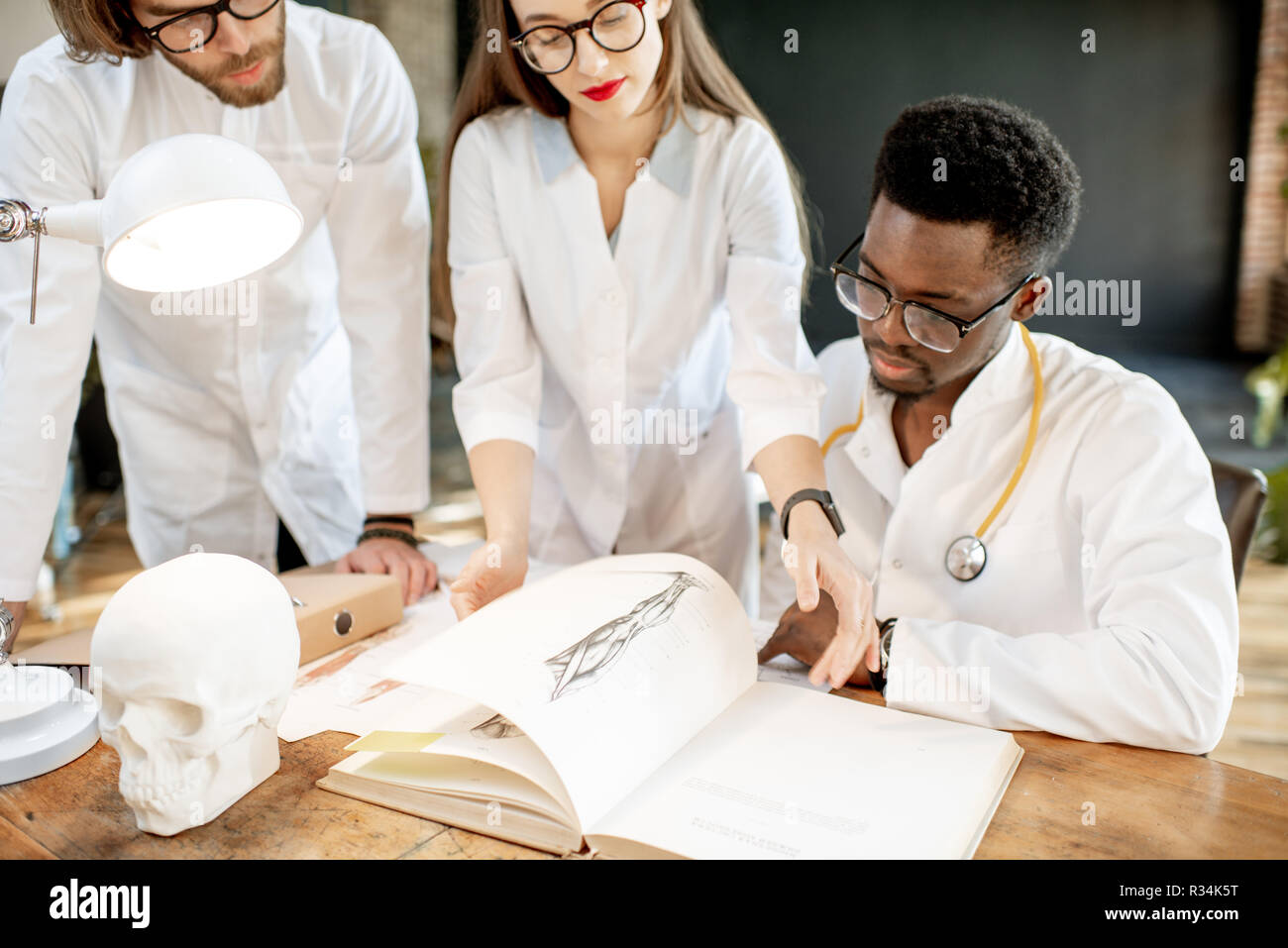 Gruppe von einem Jungen multi Ethnizität Ärzte oder Medizinstudenten in Uniform arbeiten mit Buch und Zeichnungen auf dem Tisch in der retro Office Interior Stockfoto