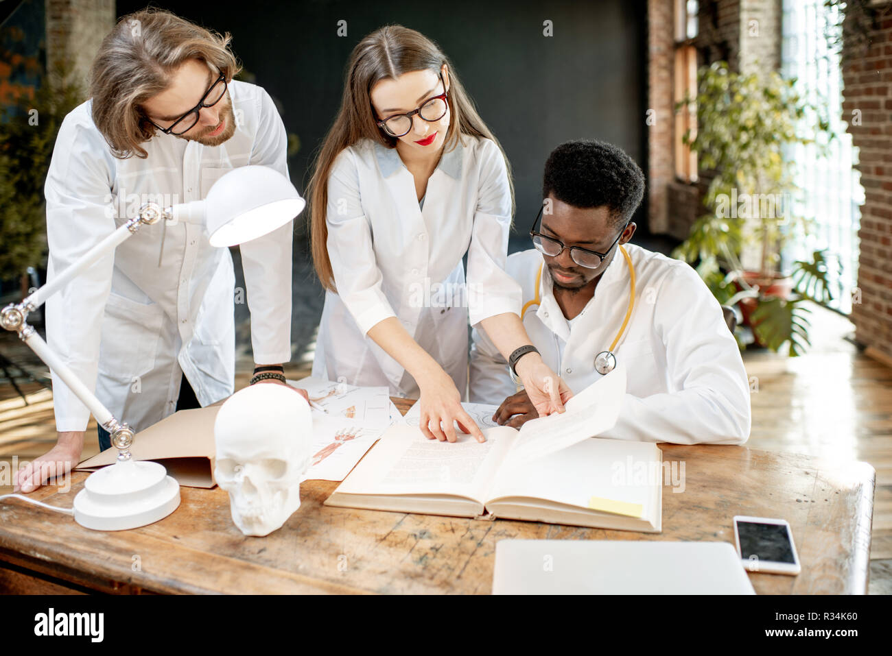 Gruppe von einem Jungen multi Ethnizität Ärzte oder Medizinstudenten in Uniform arbeiten mit Buch und Zeichnungen auf dem Tisch in der retro Office Interior Stockfoto