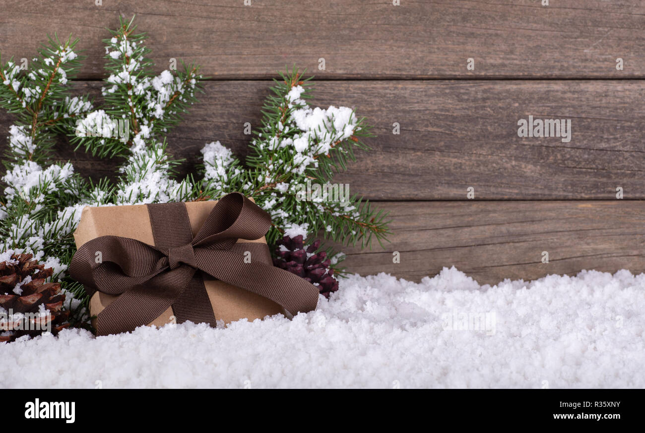 Weihnachtsgeschenk in braunes Papier und Farbband mit Tannenzapfen und immergrünen Zweigen auf Schnee gegen einen rustikalen Holzmöbeln Hintergrund gewickelt Stockfoto