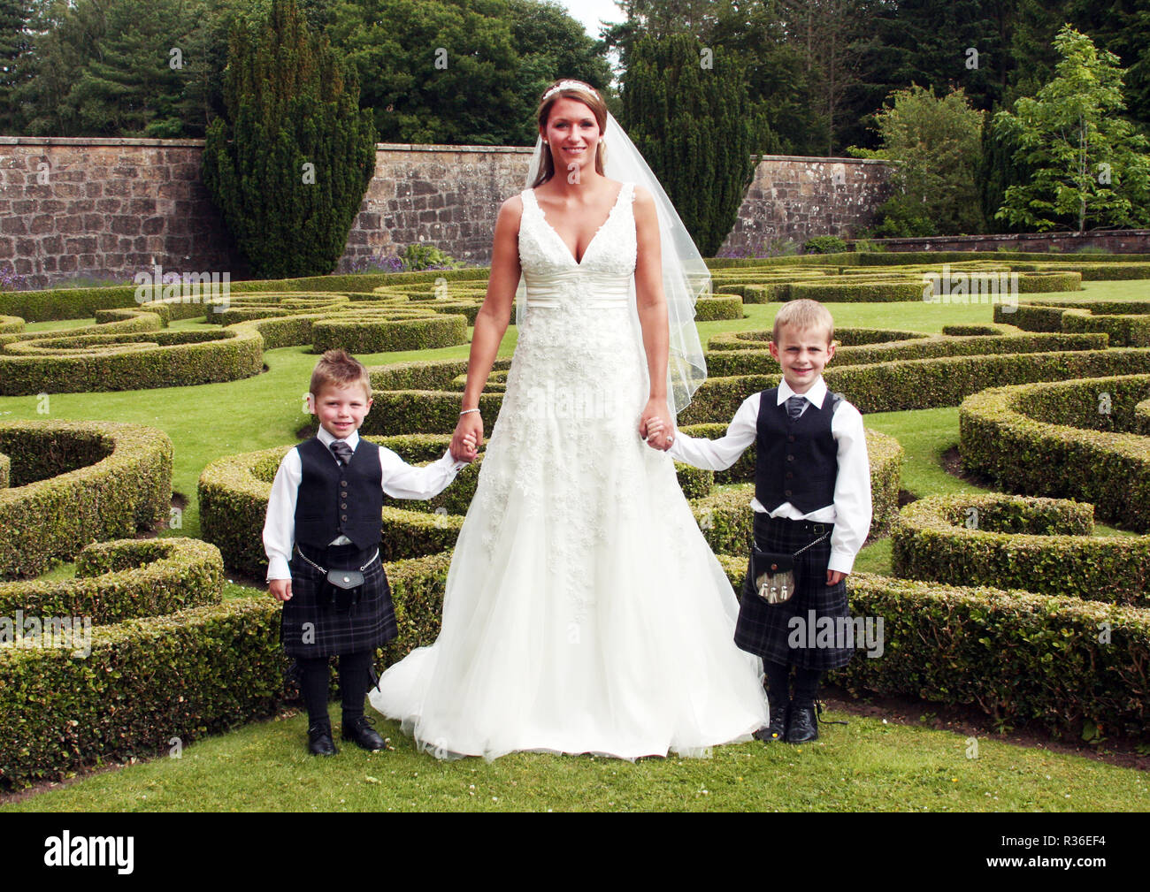 Eine Umwerfende Braut Die In Ihrem Weissen Hochzeitskleid Wirft Mit Ihren Zwei Seite Jungs In Tartan Und Kilts In Einem Mini Labyrinth Garten Nach Ihrer Hochzeit In Schottland Stockfotografie Alamy