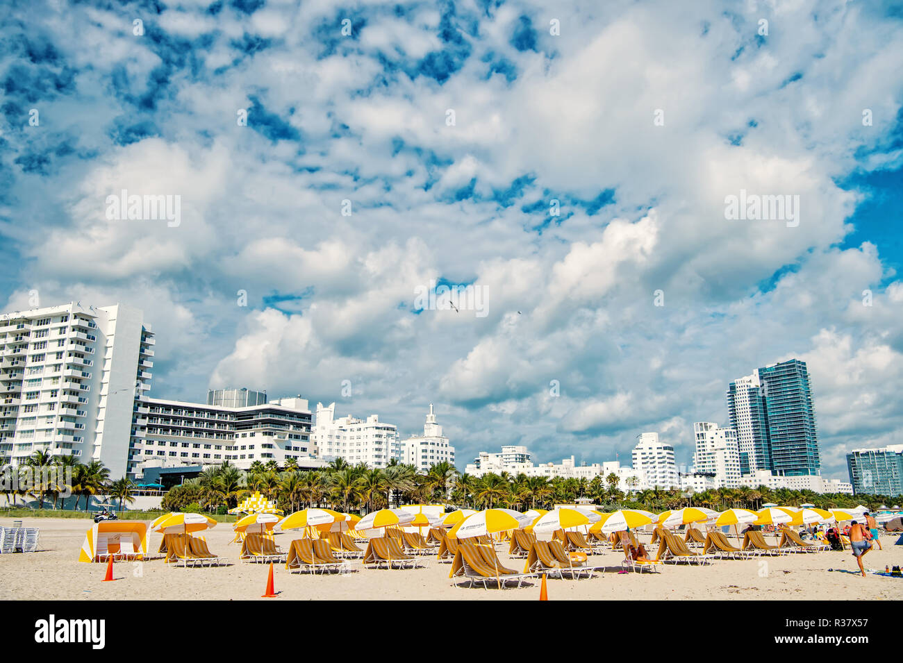 Miami, South Beach, der Strand mit Liegestühlen unter der gelben Sonnenschirmen, grüne Palmen und Mehrfamilienhäuser, Hotels oder sonnigen Tag. Die Skyline der Stadt mit weißen Wolken am blauen Himmel. Sommer Urlaub. Städtische Landschaft Stockfoto