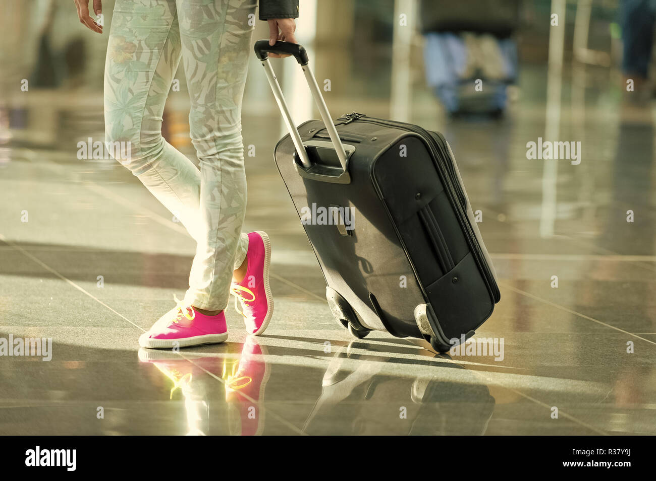 Schwarze Reisetasche oder Koffer auf Rädern in der Hand und weibliche Beine in Jeans und leuchtend rosa Gumshoes auf Füße auf grauem Fliesenboden. Reisen und Gepäck. Ferienhäuser und Mode Stockfoto