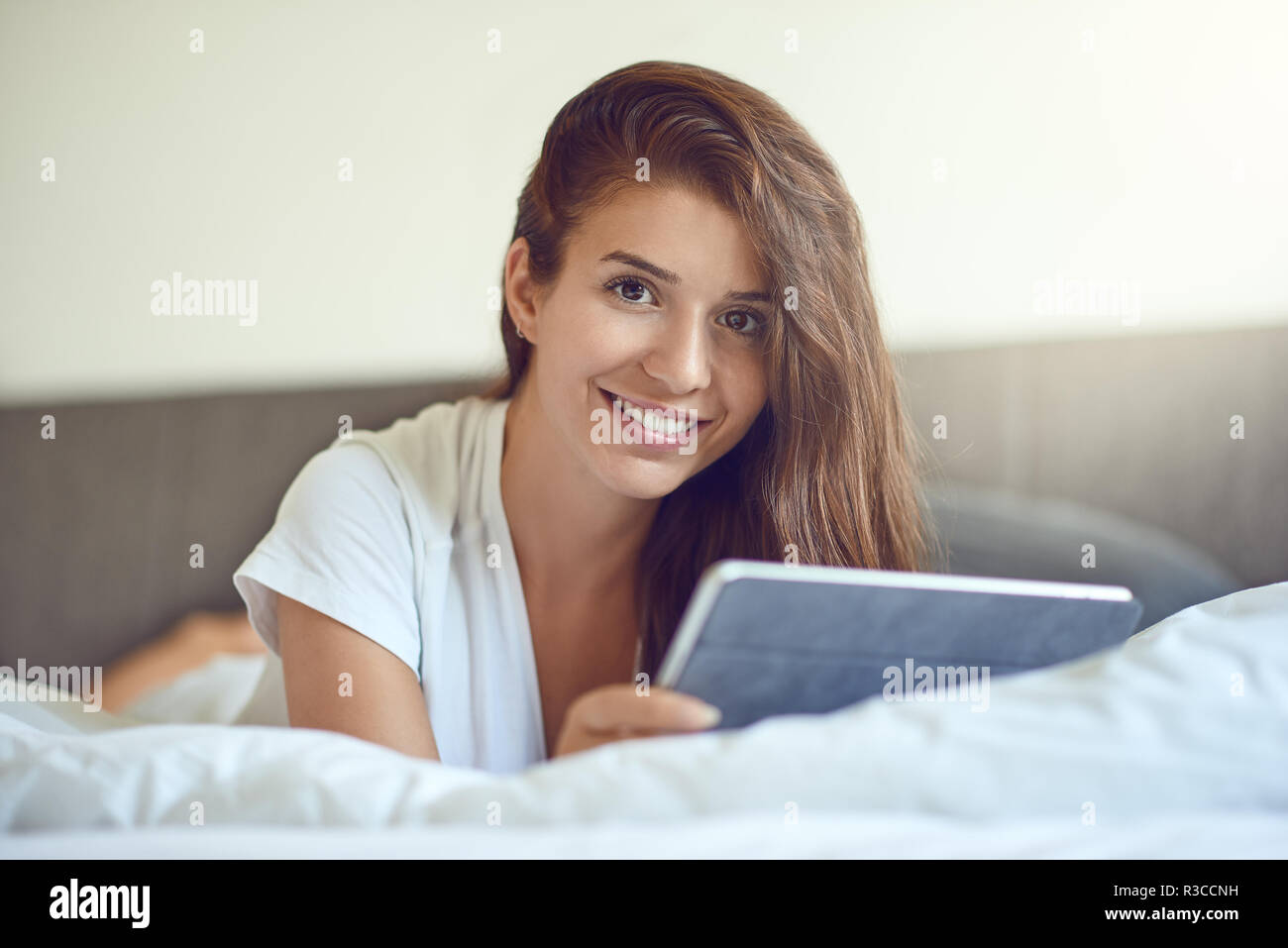 Hübsche, junge Frau mit langen braunen Haaren, Verlegung auf Magen im Bett mit weißer Bettwäsche und mit tablet pc, lächelnd in die Kamera mit Kopie Raum Stockfoto