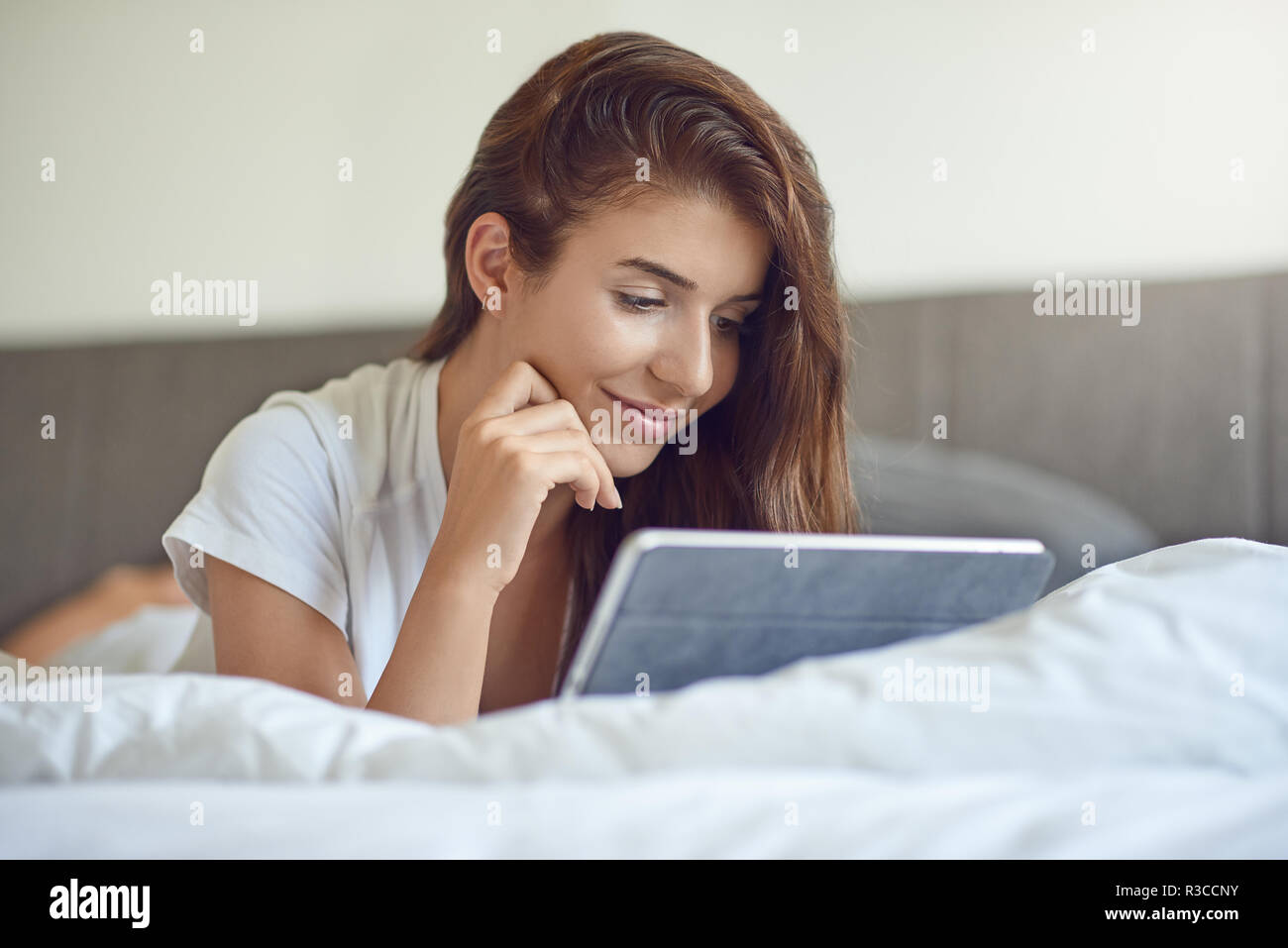 Hübsche, junge Frau mit langen braunen Haaren, Verlegung auf Magen im Bett mit weißer Bettwäsche und mit Tablet PC, Home Shopping oder beim Lesen einer E-Mail Stockfoto