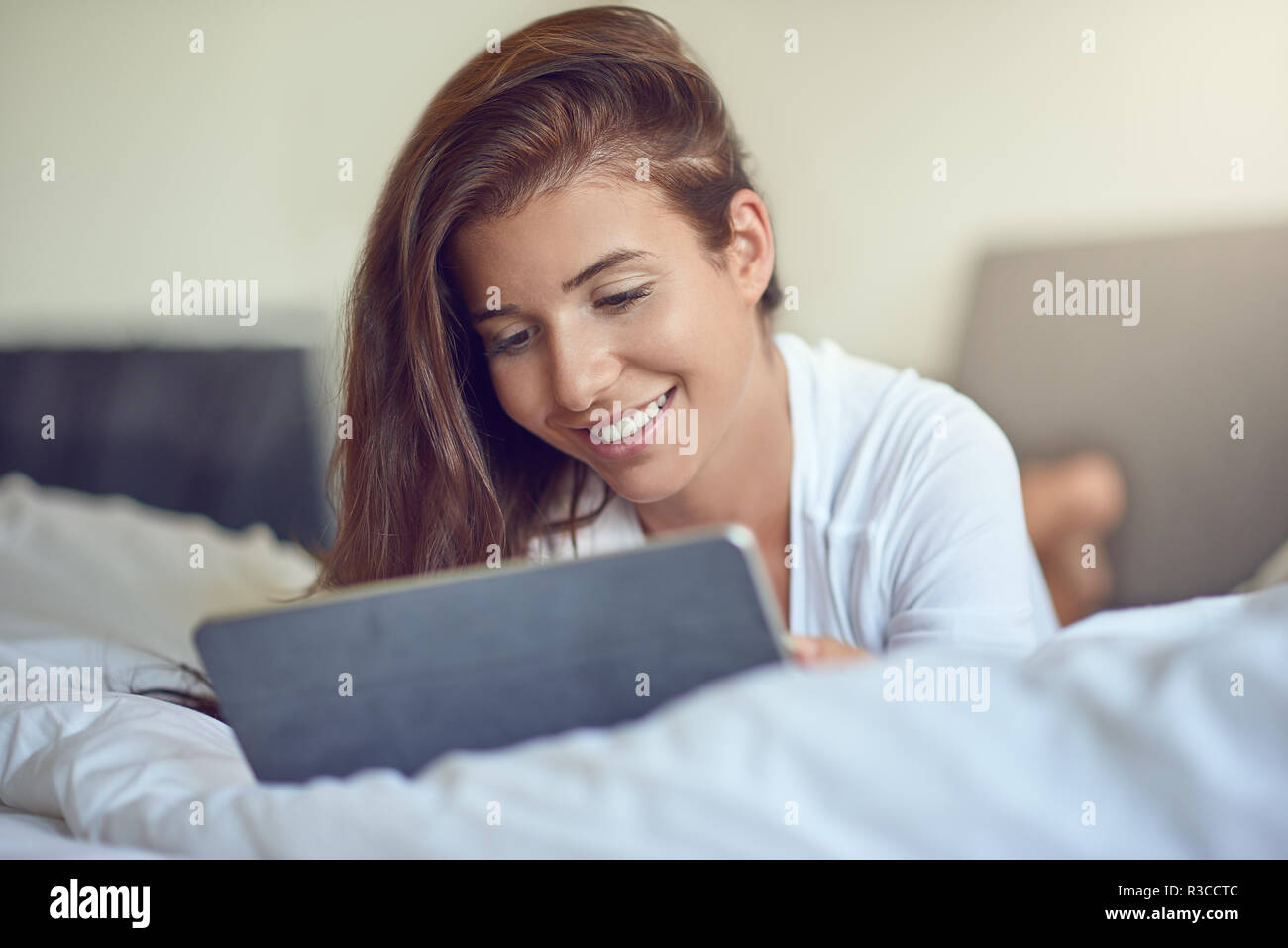 Hübsche, junge Frau mit langen braunen Haaren, Verlegung auf Magen im Bett mit weißer Bettwäsche und mit Tablet PC, Home Shopping oder beim Lesen einer E-Mail Stockfoto