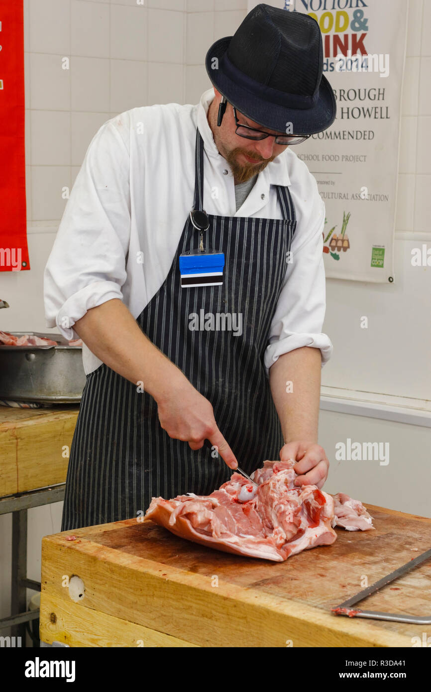 Metzger einen Hut tragen, vertikale abgestreift Schürze und einem Weißen mit einem scharfen Messer Metzger Mantel ein gemeinsames von rohem Fleisch. England, Großbritannien Stockfoto