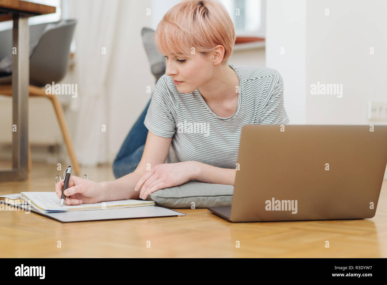 Attraktive Frau an einem Parkettboden auf einem Laptop Computer das Schreiben von Notizen in ein Notebook, wie sie von zu Hause aus oder Studien in einem Low Angle View funktioniert Stockfoto
