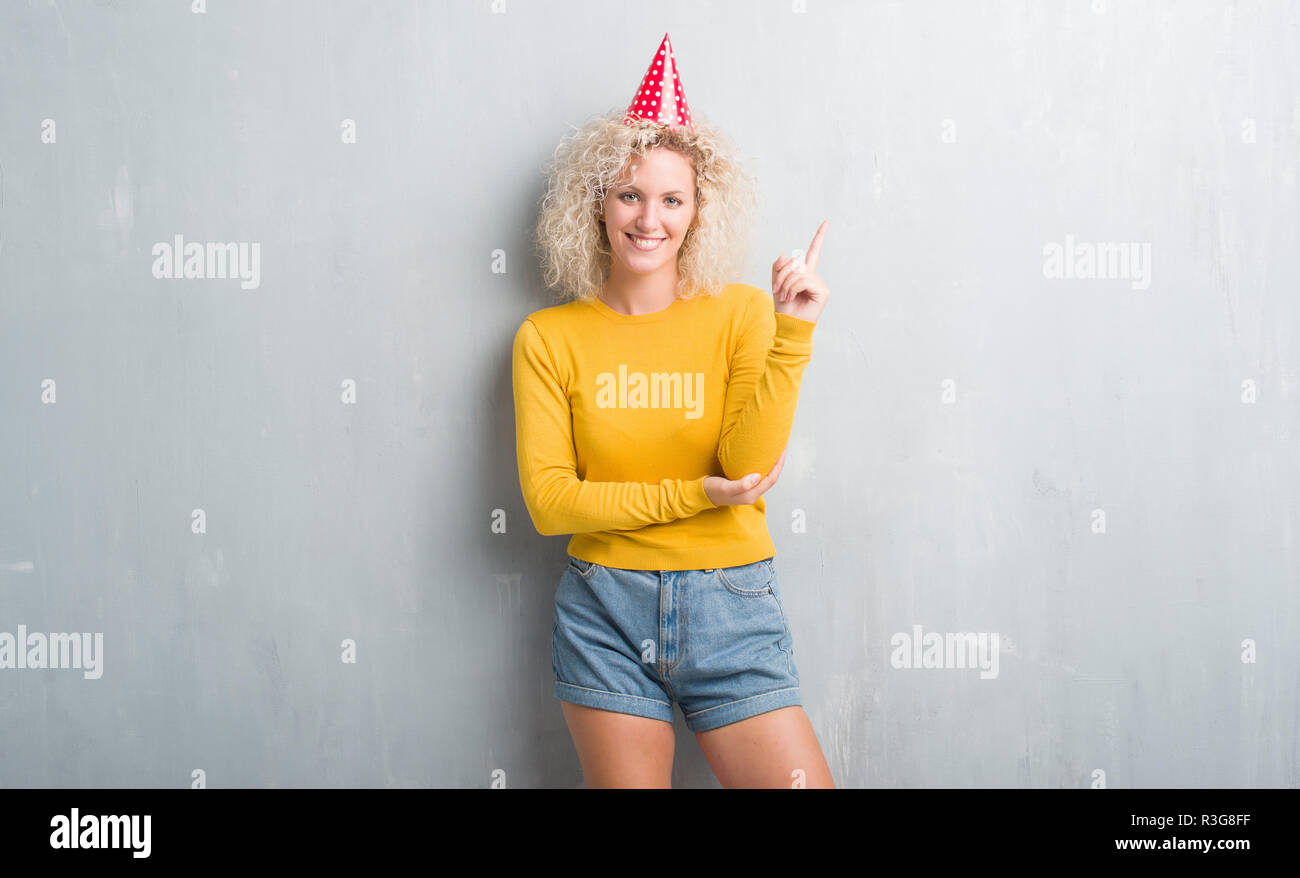 Junge blonde Frau über Grunge graue Wand tragen Geburtstag Hut mit einem Lächeln auf dem Gesicht ...
