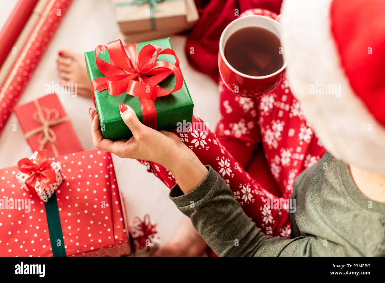 Junge Frau mit Weihnachtsmütze und xmas Schlafanzug auf dem Boden sitzend unter aufgewickelte Weihnachtsgeschenke, trinken Punsch. Über die Schulter sehen. Stockfoto