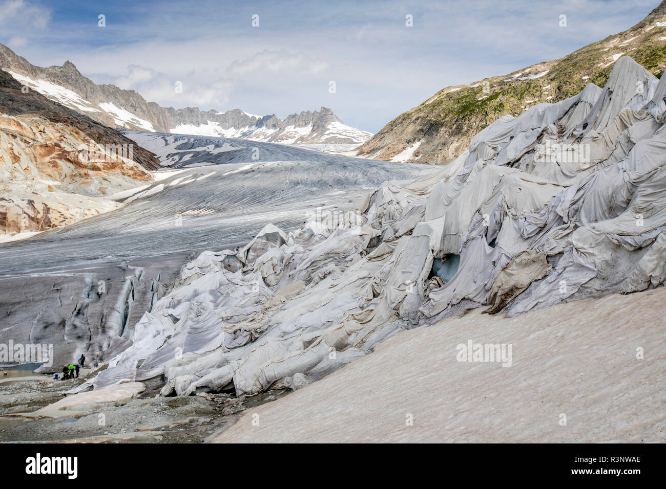 Touristen am Rhonegletscher in der Schweiz. Nach einem Winter mit viel Schnee, den meisten war es weg, wenn diese imae am 14. Juli 2018 getroffen wurde, so dass der dunkler Eis. Während Schnee eine brillante Reflektor ist der Energie von der Sonne, der dunkler Eis nimmt die Energie statt, beschleunigt das Abschmelzen der Gletscher. Die Farbe und der Dunkelheit von Gletschereis variieren in der ganzen Welt, je nach Aufbau der Verschmutzung, Alter des Eises, Partikel vom Eis und durch Mikroorganismen im Eis. Das gletschereis ist jedoch selten weiß wie Schnee. Mit kürzeren Wintern und verschwindende Schnee Cove Stockfoto
