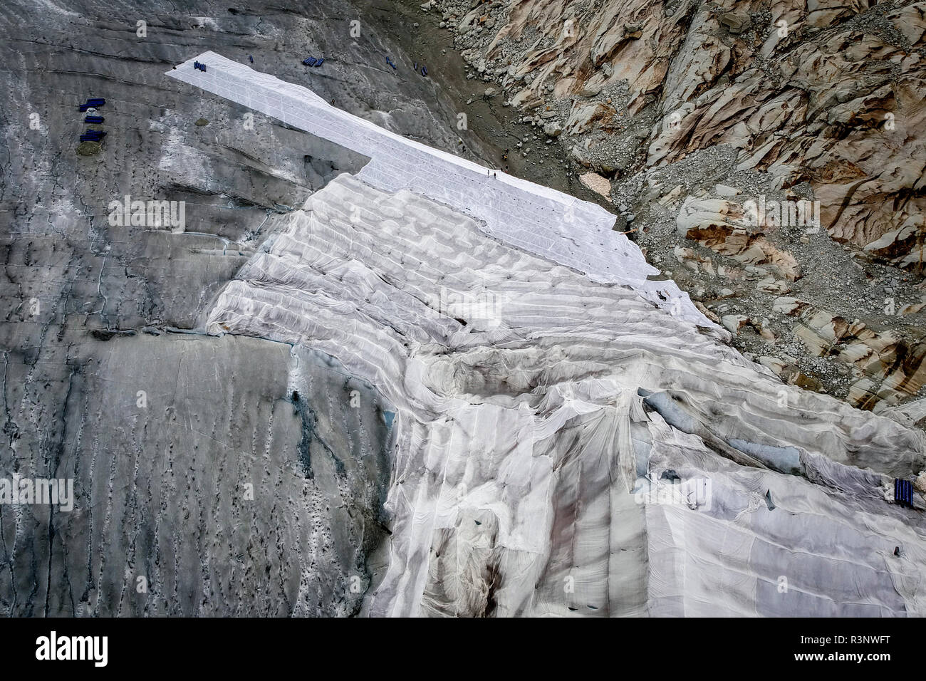Riesige Laken mit Fleece-Decken bedecken einen Eistunnel an der Mündung des Rhonegletschers in der Schweiz. Nach einem Winter mit Rekordschneemengen war der größte Teil davon verschwunden, als dieser Imae am 14. Juli 2018 aufgenommen wurde, um das dunklere Eis freizulegen. Während Schnee ein brillanter Reflektor der Sonnenenergie ist, absorbiert das dunklere Eis stattdessen die Energie und beschleunigt so das Schmelzen des Gletschers. Die Farbe und Dunkelheit des Gletschereises variiert weltweit, je nach Verschmutzungsbildung, Alter des Eises, vom Eis aufgepflückten Partikeln und Mikroorganismen im Eis. Das Gletschereis ist jedoch selten wh Stockfoto