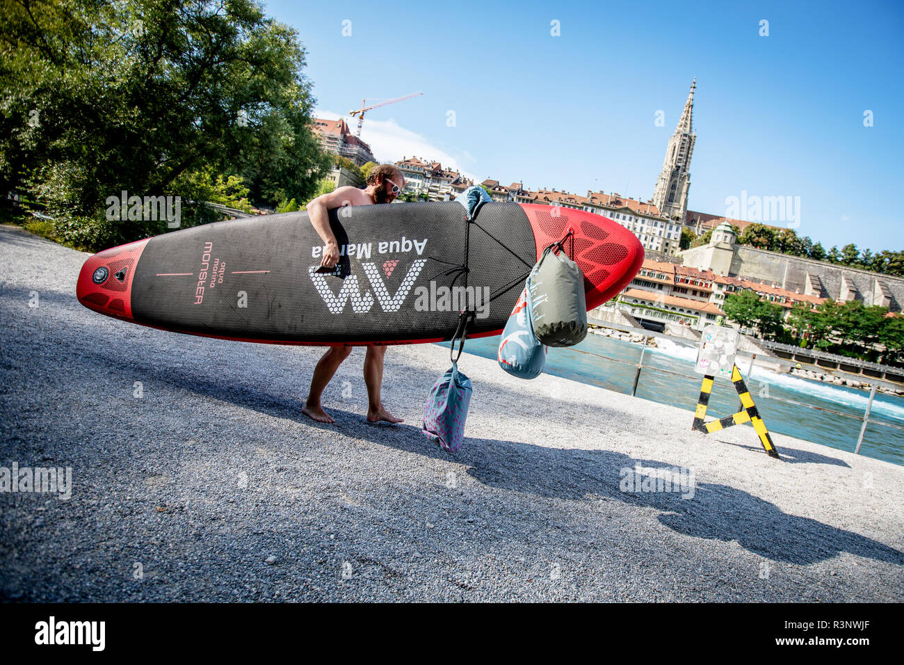 Ein Aare-Schwimmer macht sich bereit, mit einem Surfbrett und drei wasserdichten schwimmenden Aare-Taschen den Fluss Aare zu betreten. Die Aare schwebt direkt durch die Schweizer Hauptstadt Bern, und sobald die Temperatur über 10 Grad kriecht, sieht man, wie die Menschen zum Schwimmen ins Wasser springen. Viele nutzen den Fluss regelmäßig, um zum Mittagessen oder zur Arbeit zu schwimmen. Einige schwimmen sogar das ganze Jahr über. Stockfoto