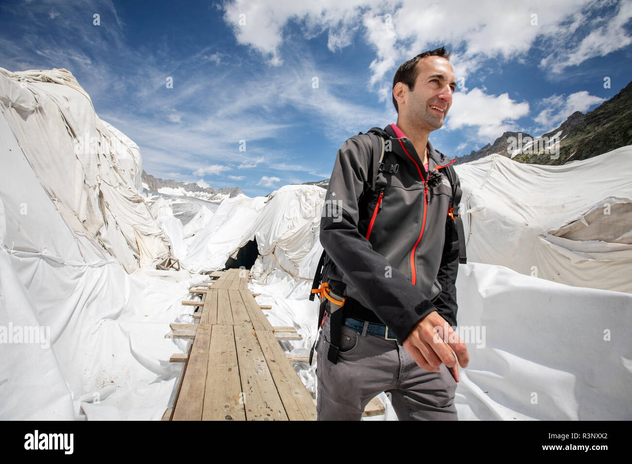 Glaciolog David Volken an der Mündung des Rhone-Gletschers. Riesige Laken mit Fleece-Decken, die einen Eistunnel an der Mündung des Rhonegletschers in der Schweiz bedecken. Nach einem Winter mit Rekordschneemengen war der größte Teil davon verschwunden, als dieser Imae am 14. Juli 2018 aufgenommen wurde, um das dunklere Eis freizulegen. Während Schnee ein brillanter Reflektor der Sonnenenergie ist, absorbiert das dunklere Eis stattdessen die Energie und beschleunigt so das Schmelzen des Gletschers. Die Farbe und Dunkelheit des Gletschereises variiert weltweit, je nach Verschmutzungsbildung, Alter des Eises, vom Eis aufgepflückten Partikeln und von Stockfoto