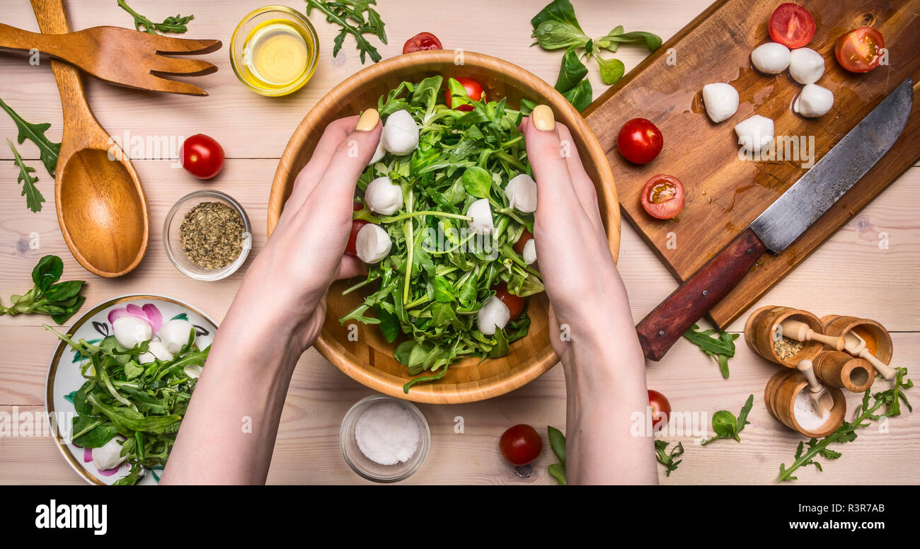 Mädchen rührt frisch zubereiteten Salat aus Rucola, Tomaten und Mozzarella mit ihren Händen in eine hölzerne Schüssel, um die Zutaten sind festgelegt Stockfoto