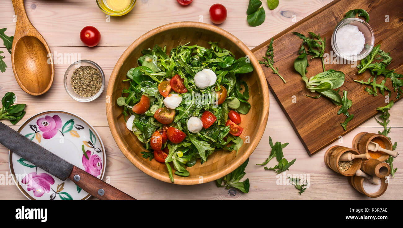 Frisch zubereiteten Salat aus Rucola, Tomaten und Mozzarella, in eine hölzerne Schüssel, durch Zutaten umgeben, rustikalen Holzmöbeln Hintergrund, Ansicht von oben Stockfoto