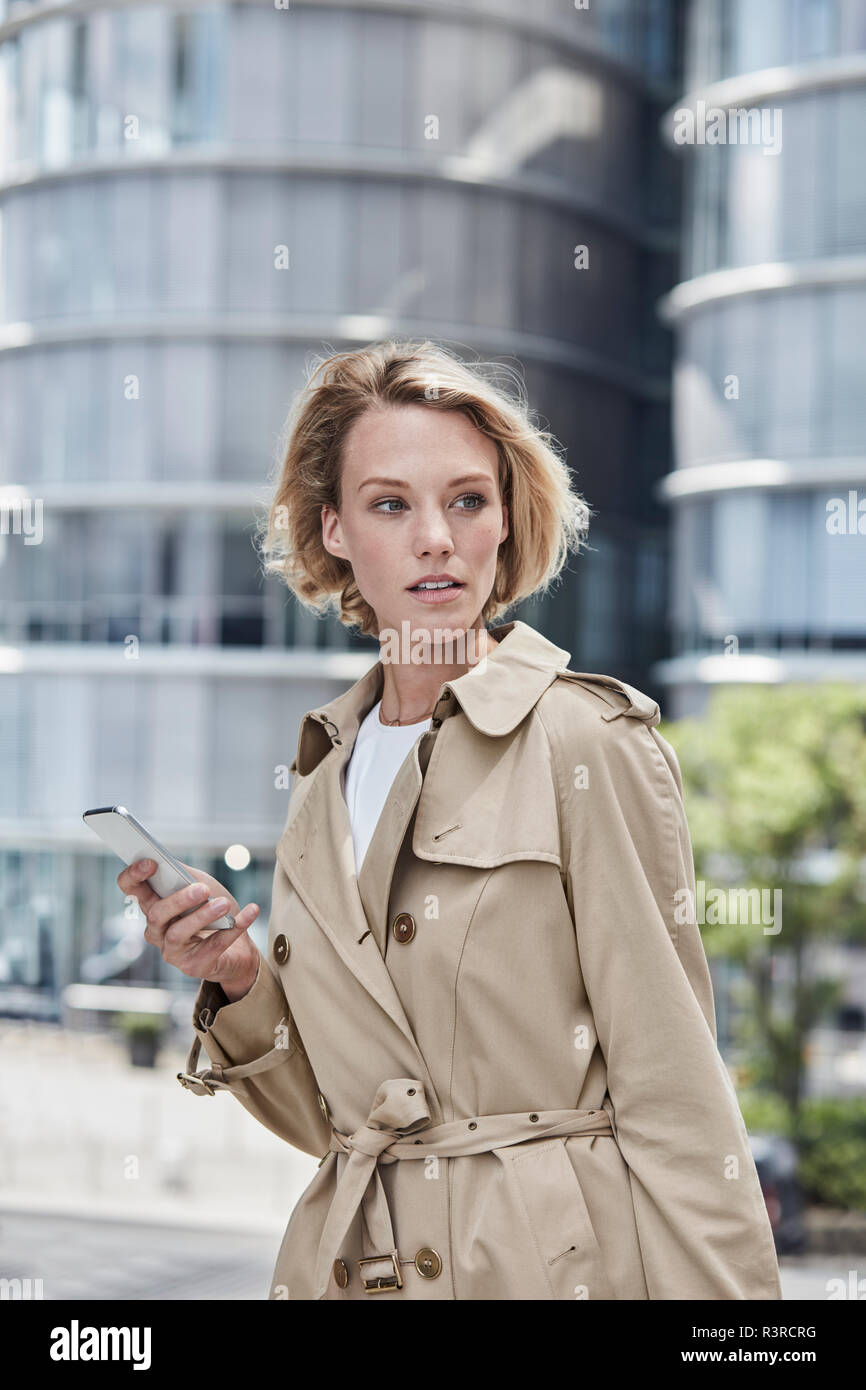 Deutschland, Duesseldorf, Porträt des blonden Jungen Geschäftsfrau mit  Smartphone tragen beige Trenchcoat Stockfotografie - Alamy