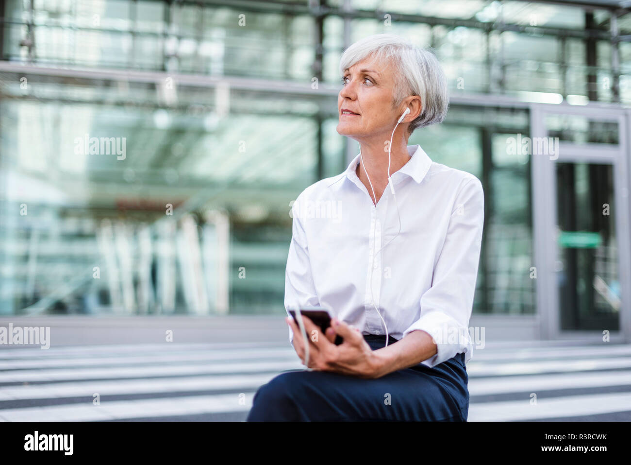 Senior Geschäftsfrau, draußen sitzen mit Smartphone und Ohrhörer Stockfoto