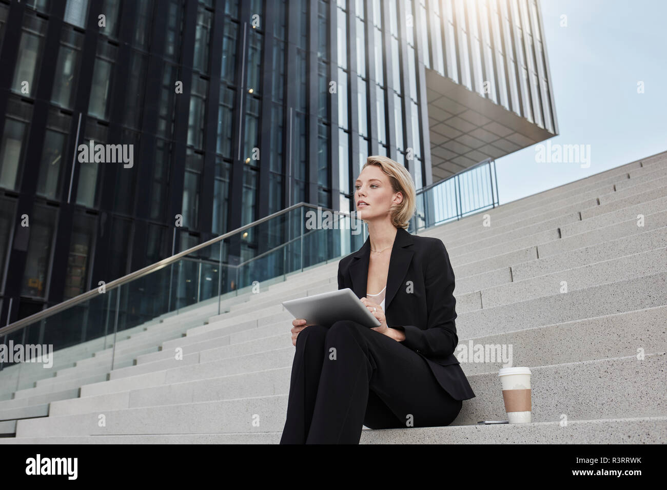 Blond Geschäftsfrau mit Tablet und Kaffee zu sitzen auf der Treppe vor der modernen Bürogebäude gehen Stockfoto