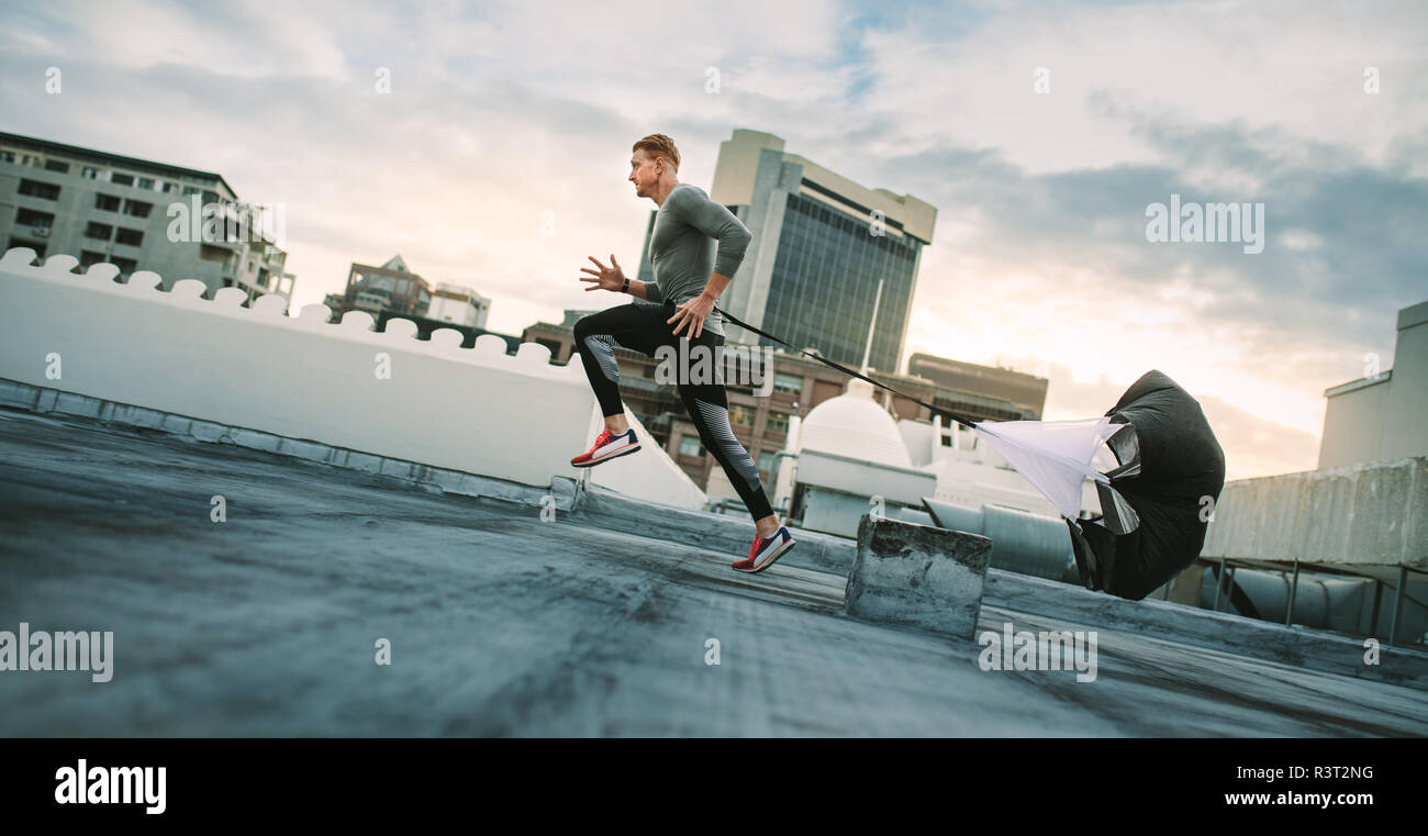 Mann tun Trainings mit Widerstand Fallschirm auf dem Dach. Seitenansicht eines Fitness Mann laufen mit einem Widerstand Fallschirm auf der Terrasse eines Gebäudes. Stockfoto
