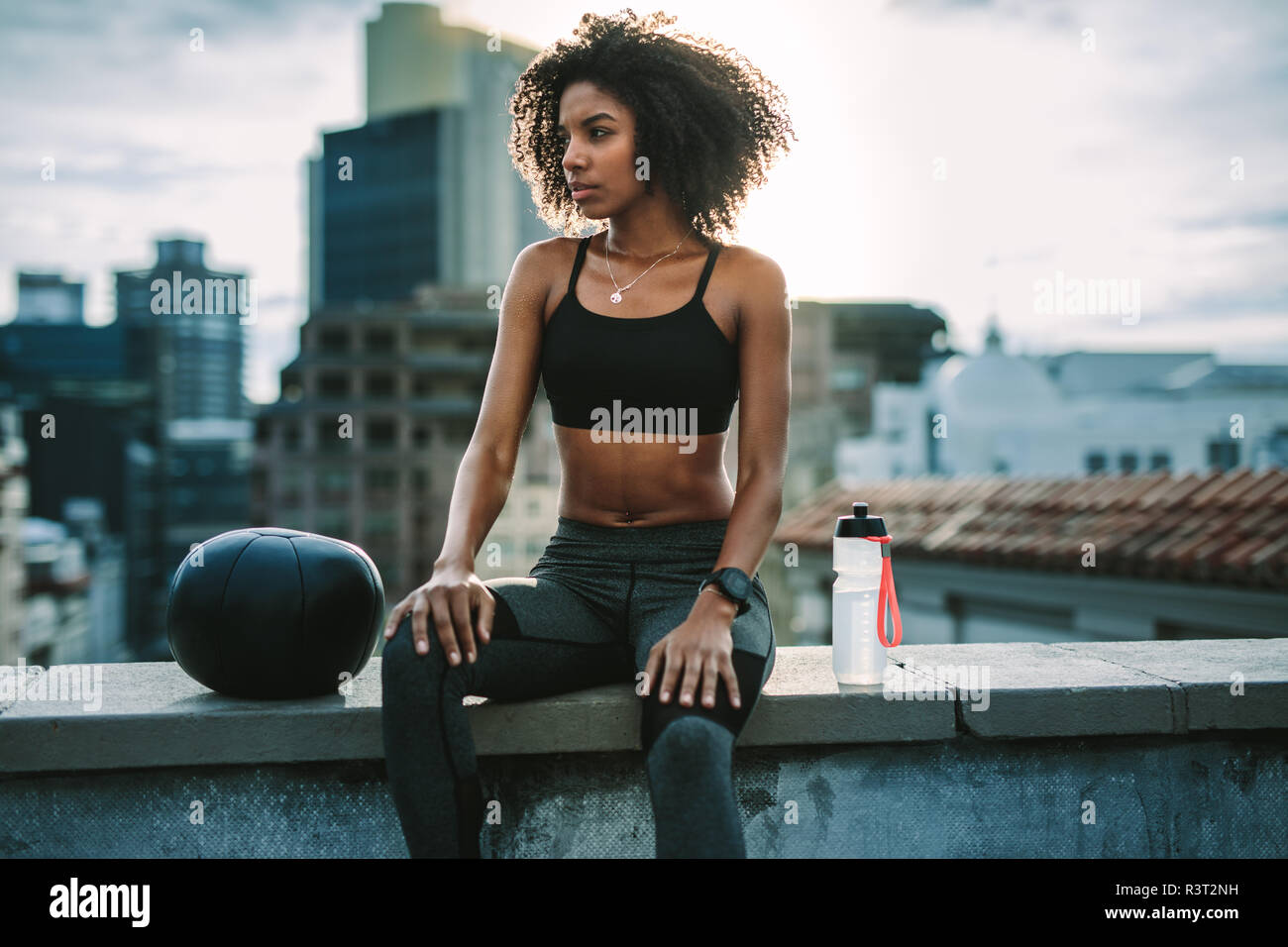 Athletische Frau sitzt auf der Dachterrasse Zaun mit einem medizinball und Wasserflasche während des Trainings. Frau in Fitness Kleidung Entspannung nach dem Training sitzen Stockfoto