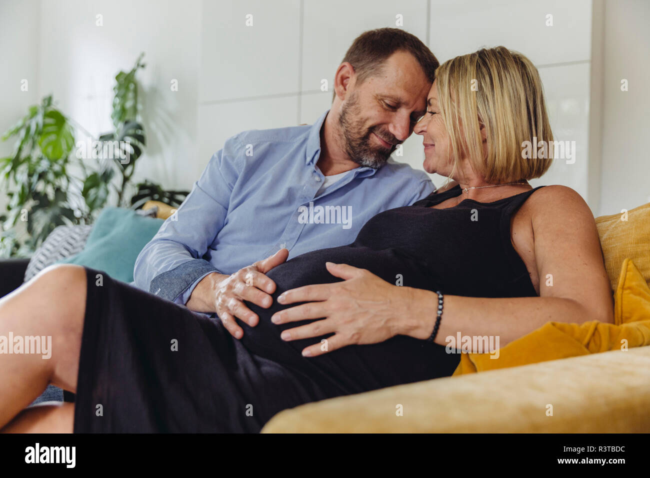 Reifer Mann und seine schwangere reife Frau sitzt auf der Couch ihren Bauch berühren. Stockfoto
