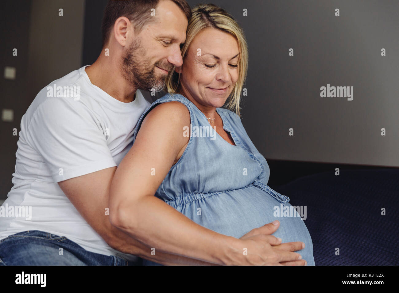 Reifer Mann und seine schwangere reife Frau sitzen auf dem Bett ihr Bauch berühren. Stockfoto