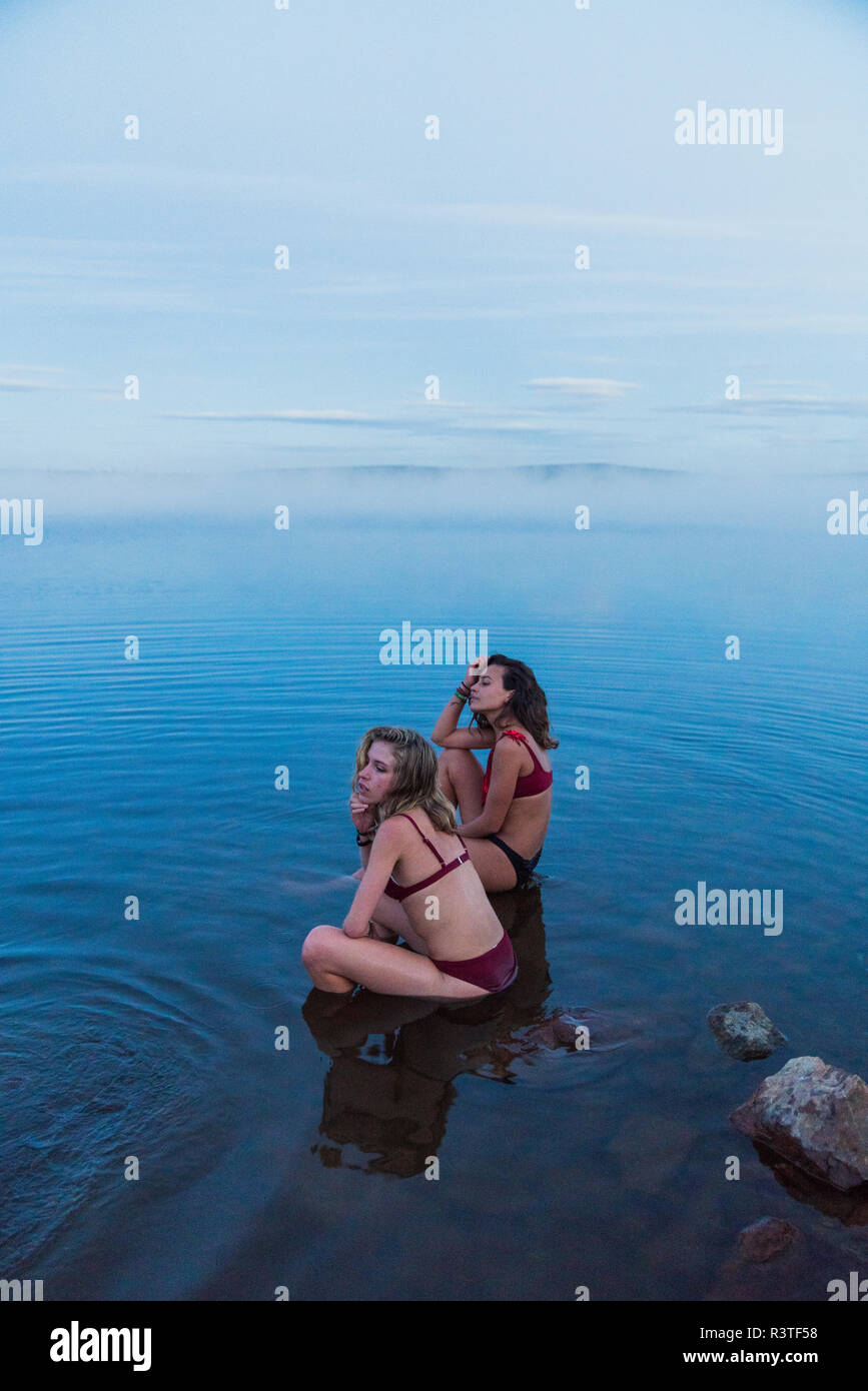 Zwei junge Frauen Bikinis tragen, sitzen auf einem Stein in einem See Stockfoto