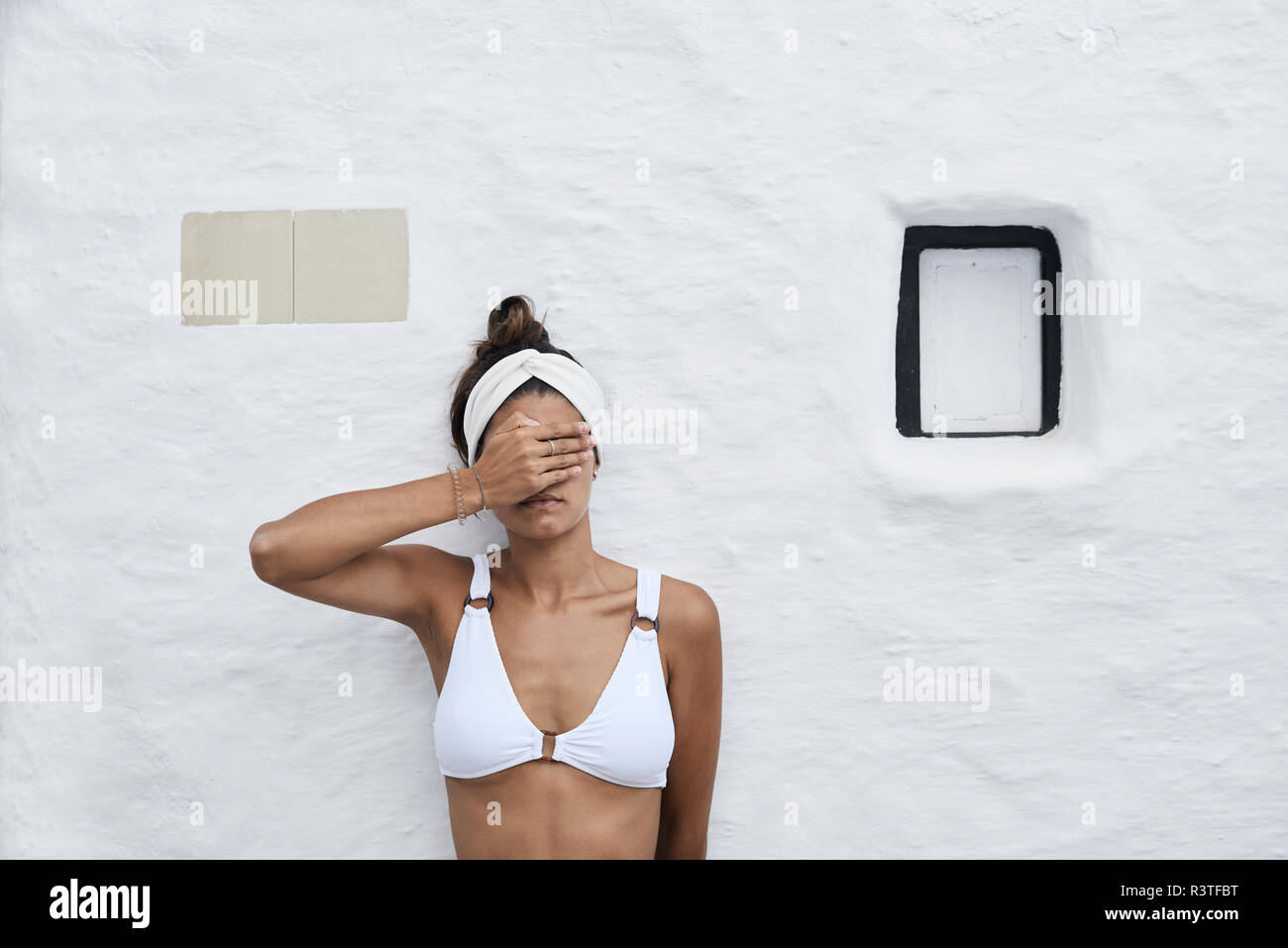 Junge Frau mit weißen Bikini Top gegen weiße Wand gelehnt, die Augen mit der Hand Stockfoto