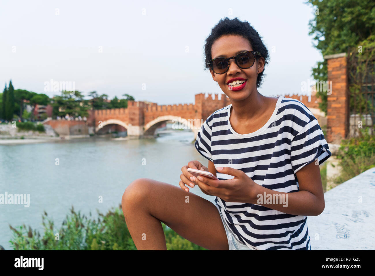 Lächelnden jungen Frau Sonnenbrille tragen im Riverside mit Handy Stockfoto