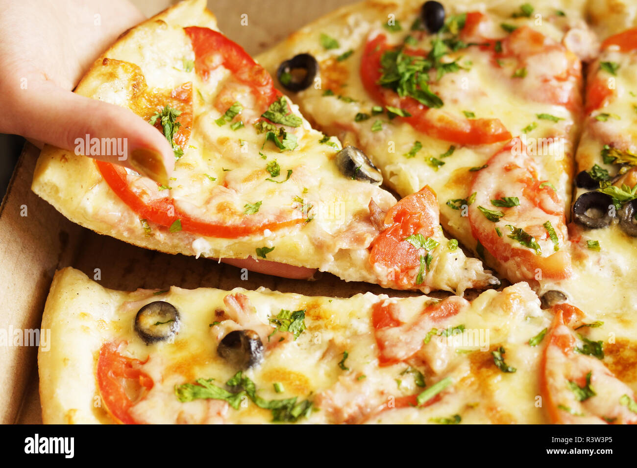 Foto der jungen Frau, die mit ihrer Hand ein Stück heiße frisch gebackene Pizza mit Käse, Schinken und Hähnchenfleisch, Tomaten und Oliven aus pizza box Stockfoto