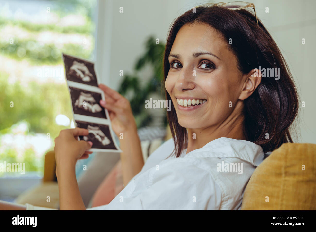 Portrait von lächelnden Frau mit Ultraschall Bilder von ungeborenen Kind auf der Couch Stockfoto