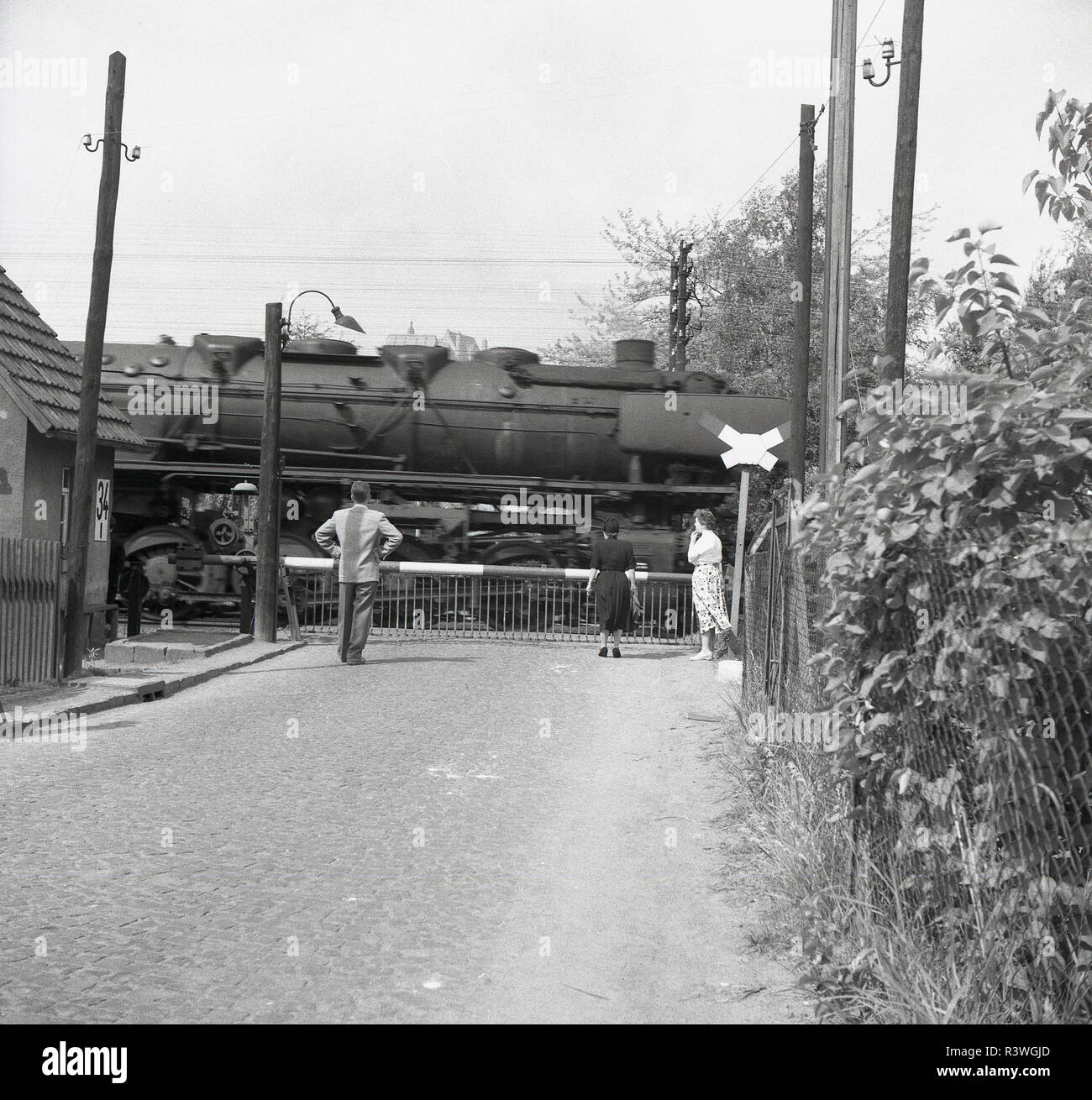 1950, Jugoslawien, Osteuropa, die Barrieren auf einer hand Bahnübergang über eine Seitenstraße betrieben werden geschlossen, wie eine große Dampflok Vergangenheit geht. Stockfoto