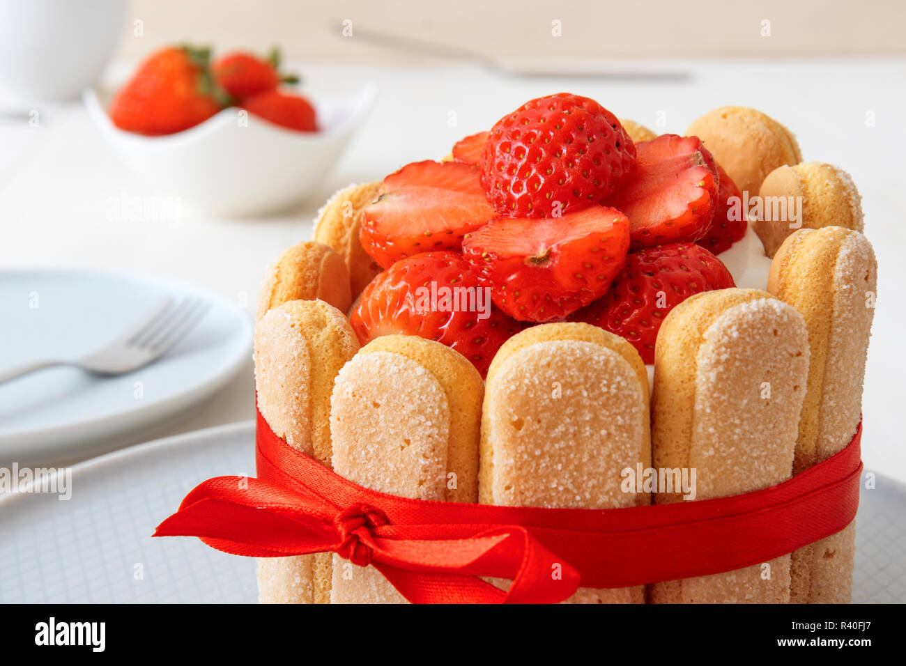 Charlotte, französische Dessert mit Erdbeeren, gefesselt mit einem roten Band, Tasse, Schüssel mit Erdbeeren auf weißer Tisch. Stockfoto