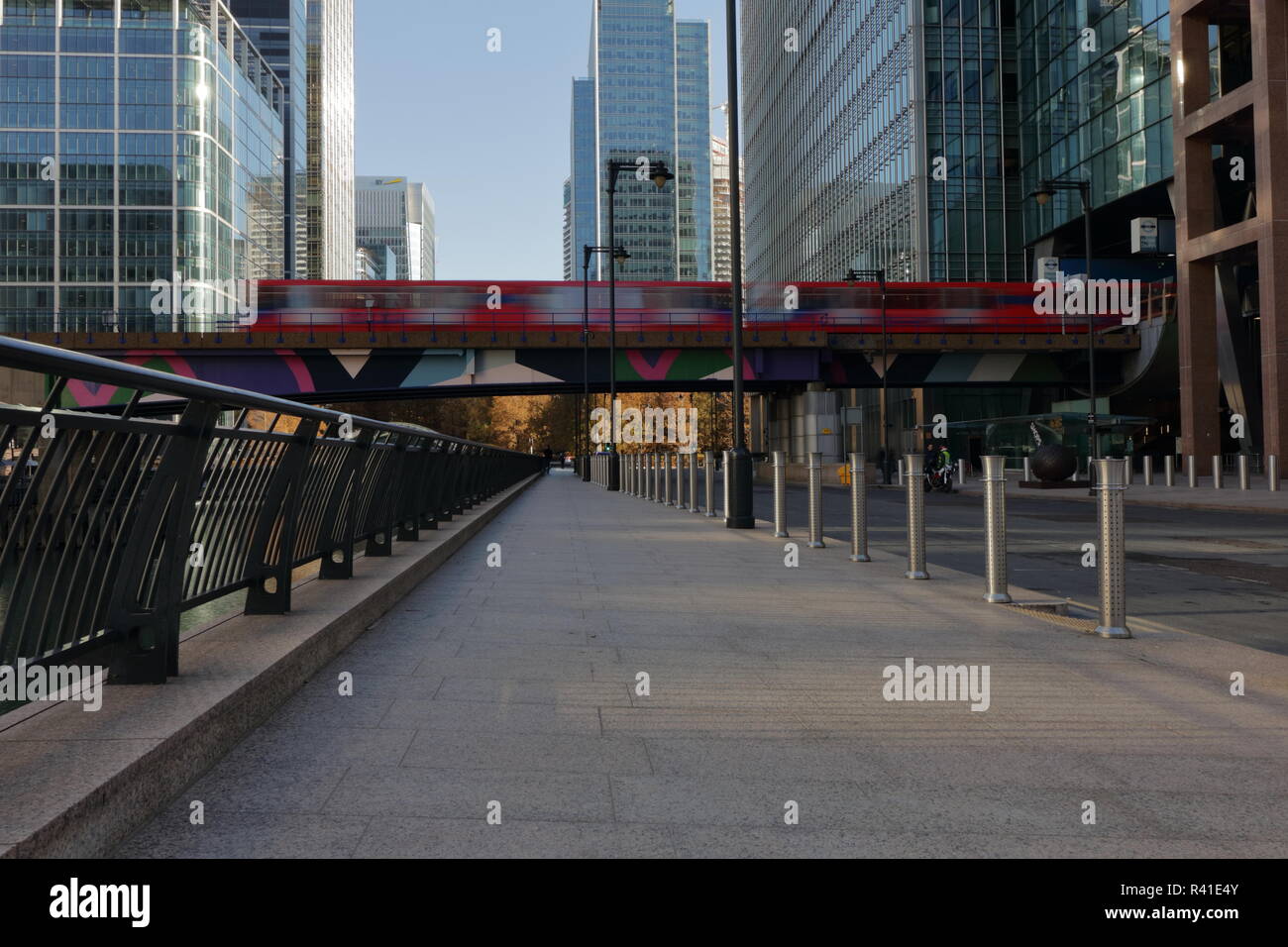 Am frühen Sonntagmorgen in Canary Wharf in London mit DLR-Zug vorbei und hinten leer Bank Street. Schönen Tag mit blauem Himmel und einige Überlegungen. Stockfoto Am frühen Sonntagmorgen in Canary Wharf in London mit DLR-Zug vorbei und hinten leer Bank Street. Schönen Tag mit blauem Himmel und einige Überlegungen. Stockfoto