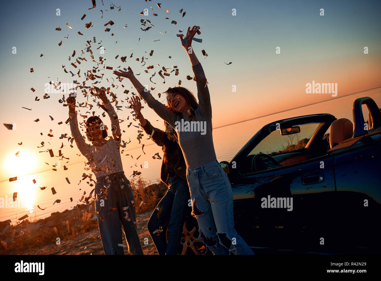 Fröhlicher junger drei Frauen tanzen in der Nähe der Cabrio. Sie werfen die fallenden Blätter. Schönen Sonnenuntergang im Hintergrund Stockfoto