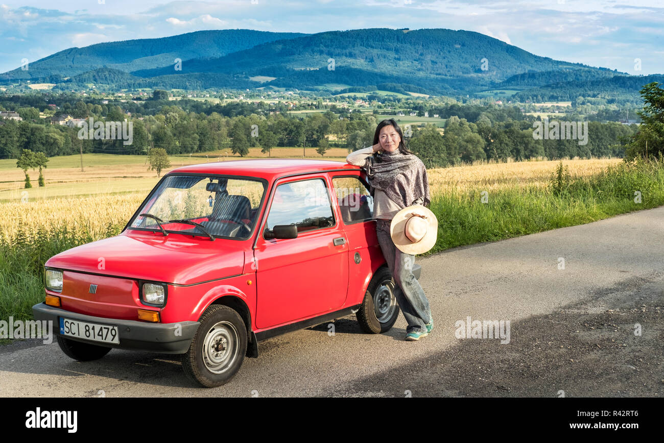 Ein Mädchen lehnte sich gegen eine rote Maluch - Polski Fiat 126p-mit Feldern und Bergen im Hintergrund Stockfoto