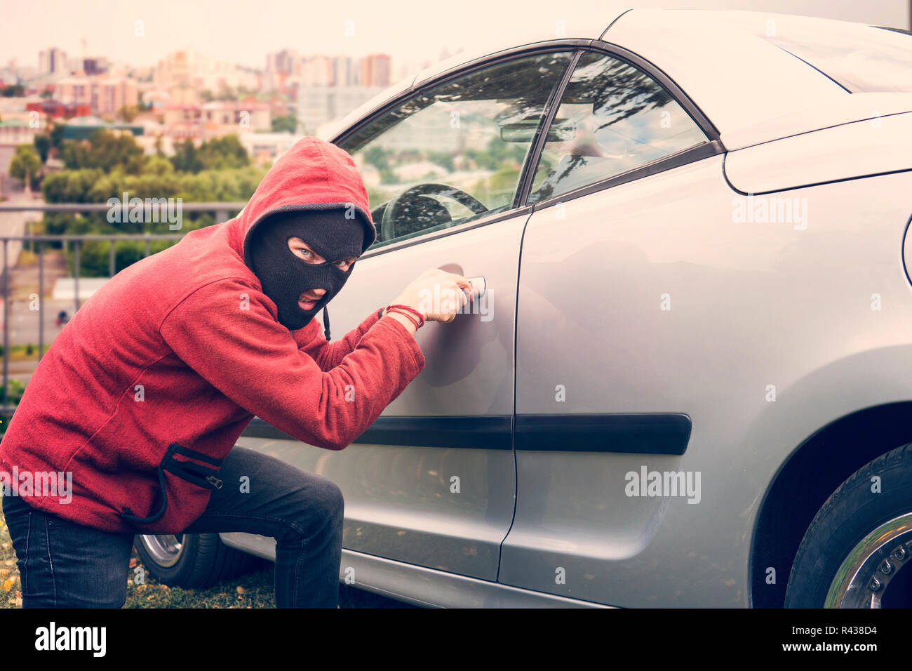 Maskierte Mann hockt und bricht ein Auto auf den Betrachter. Dieb in Hoodie und verborgene Gesicht macht versuchen, picklock das geparkte Fahrzeug zur Festsetzung Stockfoto