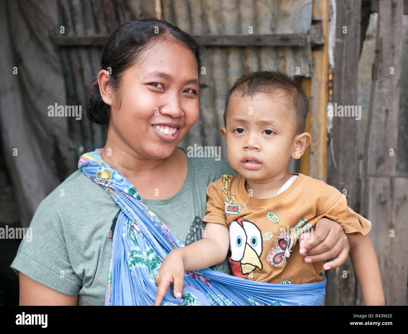 Horizontale Portrait von lächelnden, Stolz, glücklich jungen indonesischen Mutter holding Junge tragen gelbe T-shirt vor Holz und Zinn. Stockfoto