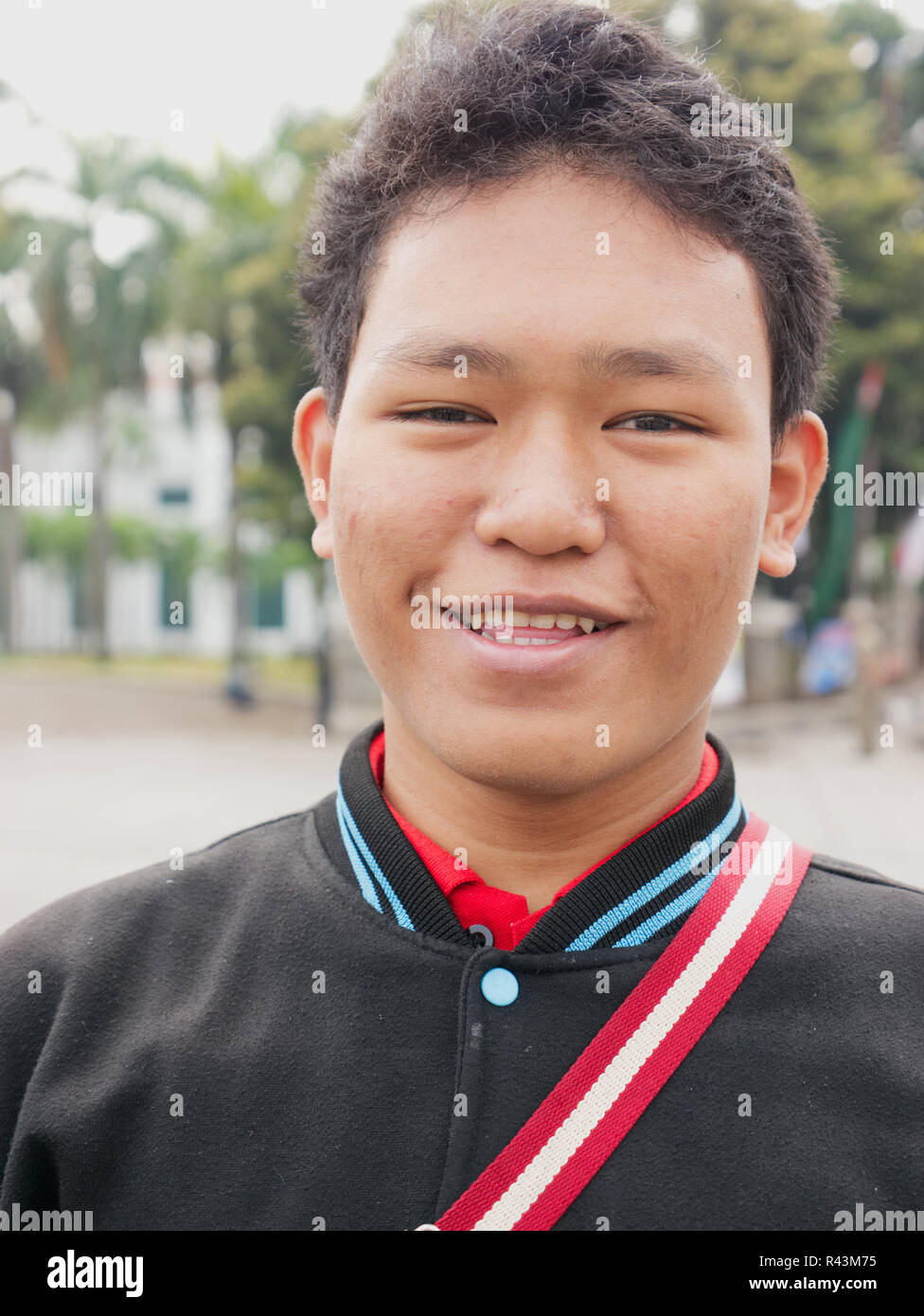 Vertikale Porträt der jungen indonesischen Teenager mit schwarzen Jacke mit roten und weißen Riemen über der Brust in die Kamera schauen und lächeln. Stockfoto