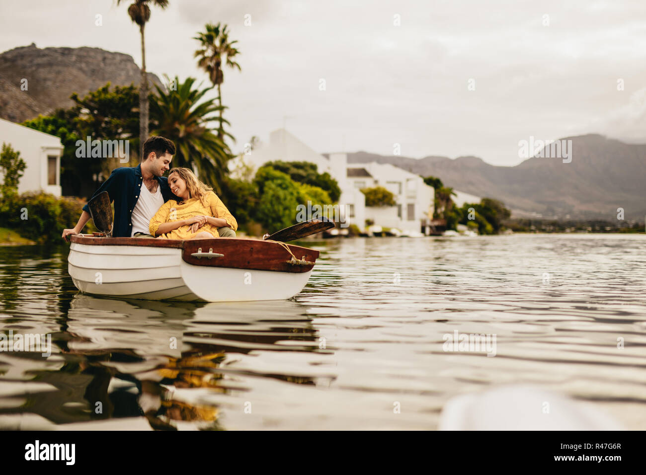 Junger Mann und Frau auf einem Boot Datum in einem See mit Hügeln und Häuser im Hintergrund. Frau sitzt entspannt lehnte sich auf ihren Freund auf einem Boot. Stockfoto